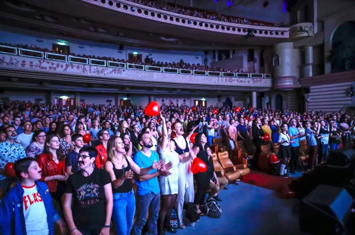 Un public nombreux à Lille se lève et applaudit au Cinéma Karaoké, certains tenant des ballons rouges, les balcons visibles au-dessus.