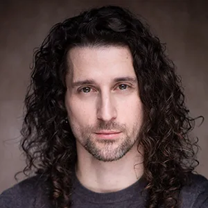 Une personne aux longs cheveux bruns bouclés et à la barbe, vêtue d'une chemise sombre, regarde directement la caméra sur un fond neutre.