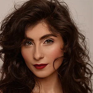 Femme aux longs cheveux noirs ondulés, rouge à lèvres rouge vif et maquillage naturel, regardant directement l'appareil photo.