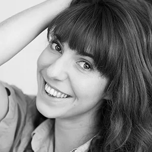 Femme aux cheveux longs et à la frange souriant à l'appareil photo, une main touchant sa tête, sur une photo en noir et blanc.