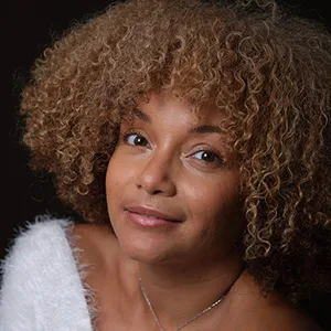 Une femme aux cheveux bruns clairs et bouclés, vêtue d'un haut blanc décolleté, regarde l'appareil photo sur un fond sombre.
