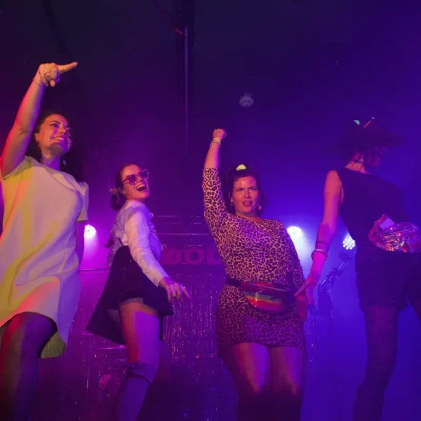 Quatre femmes posent et dansent sur une scène sous des lumières colorées, vêtues de tenues de fête éclatantes.