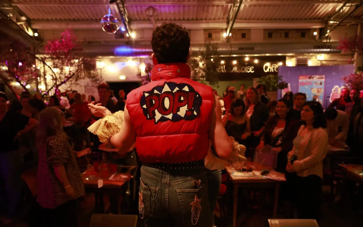 Une personne vêtue d'un gilet rouge "POP !" s'adresse à une foule assise dans une salle parisienne faiblement éclairée, canalisant l'énergie d'IDOL.
