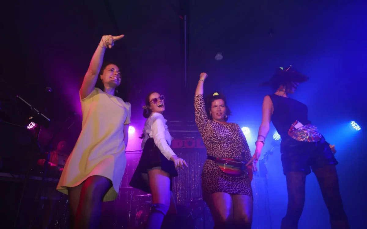 Quatre femmes posent sur scène comme des IDOLs dans des tenues rétro 80/90/2000, dansant sous des lumières colorées avec un flair parisien.